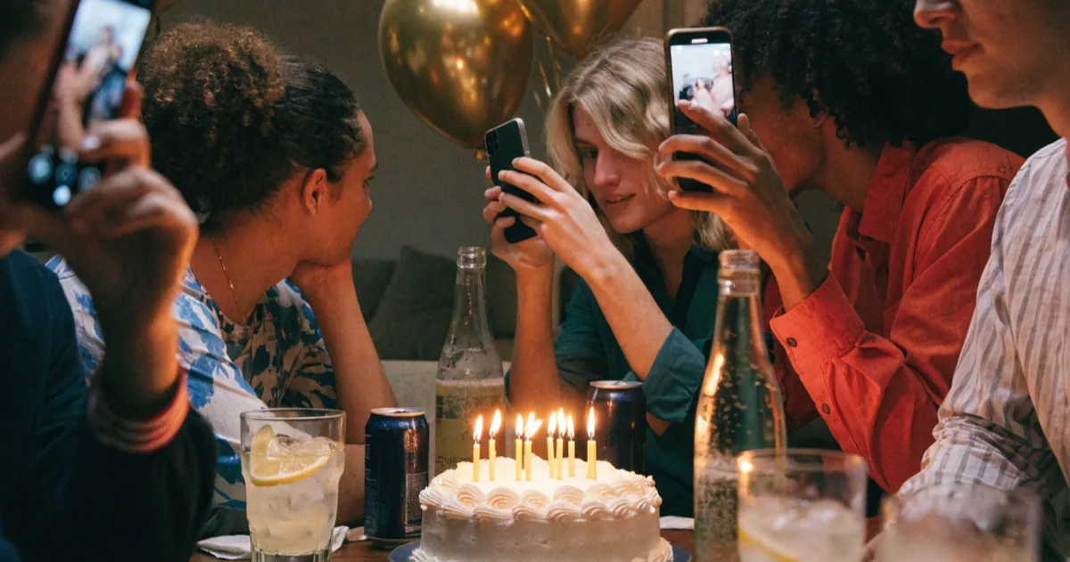 Friends crowded around a table of lit candles and gold balloons at an 18th birthday party, phones raised
