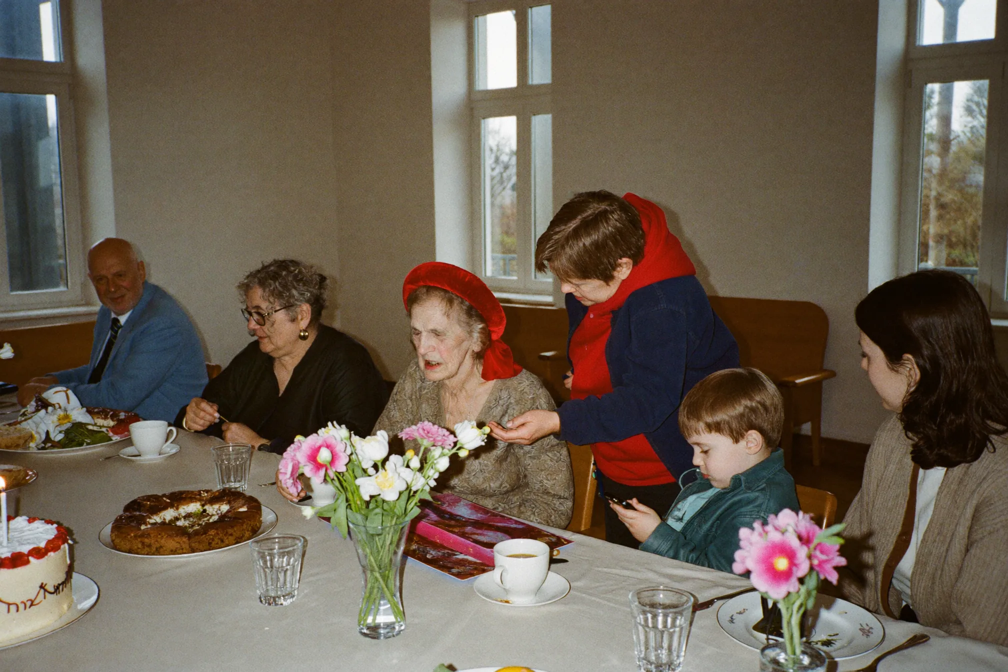 Drei Generationen um eine sitzende Frau beim hellen Mittagessen zum 90. Geburtstag, hinter ihrem Stuhl ein großer goldener Ballon in Form einer 90
