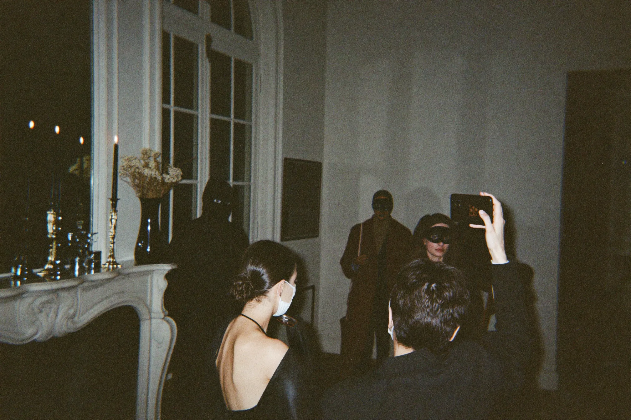 Des invités dansent lors d’une soirée pendant que l’un d’eux photographie la scène au téléphone, avec une lumière chaude qui garde les visages visibles